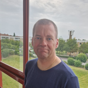 Blå ögon, kort brunt hår, man och mörkblå t-shirt. På min balkong framför glasen som är inramade i röd metal. Utanför är det ljusblå himmel och grönt gräs. Där är hus, buskar och träd i bakgrunden.
